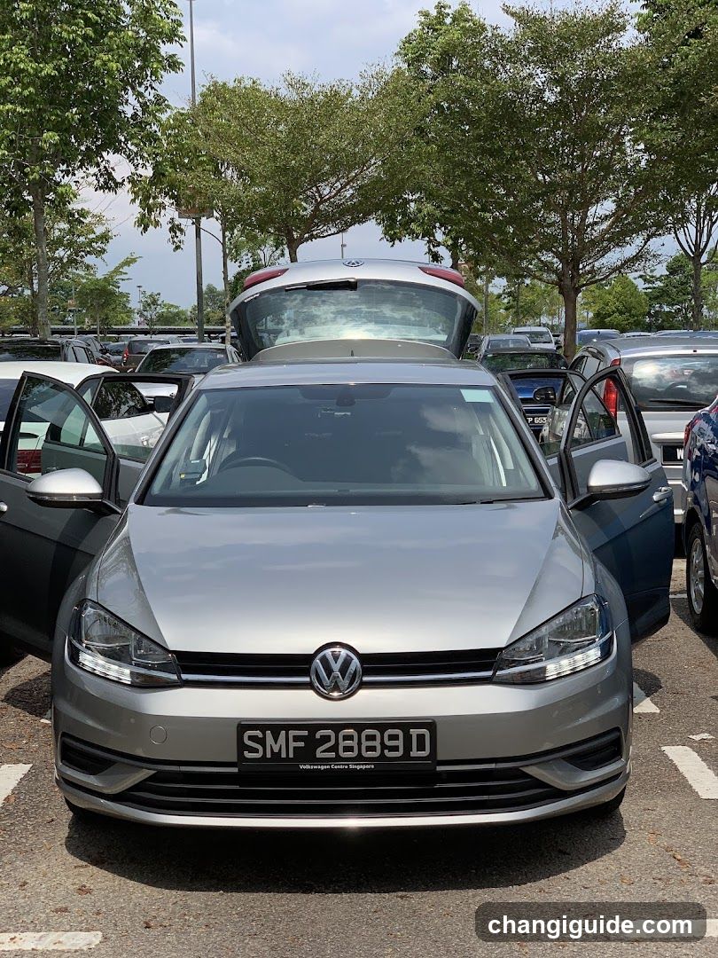 A silver Volkswagen Golf is parked in a lot with its doors open and trunk lid raised, surrounded by other cars under trees.