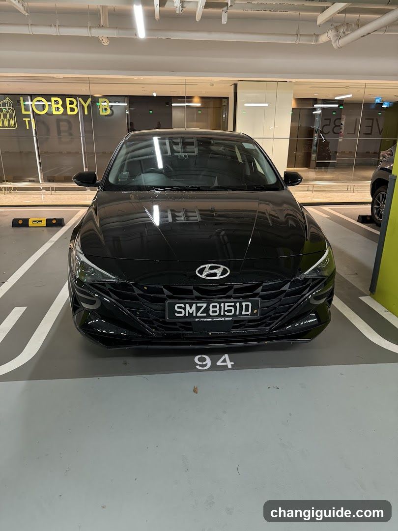 A black Hyundai sedan is parked in a garage space directly in front of a glass storefront with visible signage reading 'HOBBY' and partial text 'TI'. The shop entrance features large transparent windows reflecting the parking area, with interior lighting visible inside. A yellow sign on the left side displays an icon resembling a toy or building block. No menu boards are present.