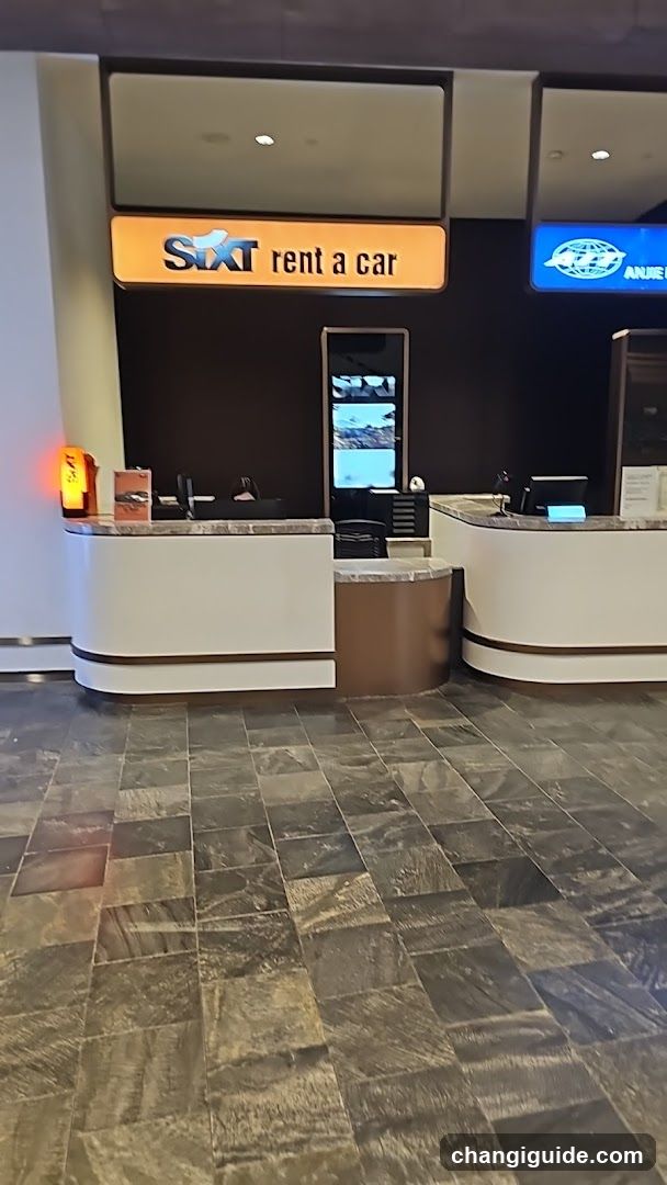 Interior view of the Sixt rent a car counter area, featuring two white reception desks with marble countertops and dark wood accents. A digital screen is mounted on the wall between counters. The floor has patterned stone tiles. An orange lamp glows on the left desk. Signage above includes 'SIXT rent a car' in yellow and blue branding.