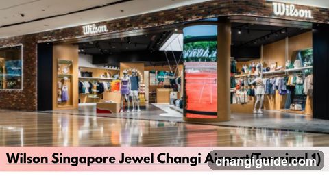 A wide-angle photo of the Wilson sports store entrance at Singapore's Jewel Changi Airport Terminal 1. The storefront features a brick facade with illuminated 'Wilson' signage above two main entrances. Inside, mannequins display clothing and accessories under bright lighting. A cylindrical digital screen displays tennis imagery near the center aisle. Shelves on the right hold folded apparel and bags. The polished floor reflects overhead lights.