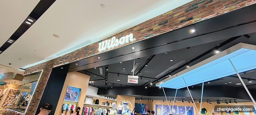 The image shows the storefront of a Wilson store, featuring a prominent 'Wilson' sign mounted on a brick facade above the entrance. The interior is visible through large glass windows and doors, displaying various sports apparel and merchandise. A blue awning extends over part of the entrance area.