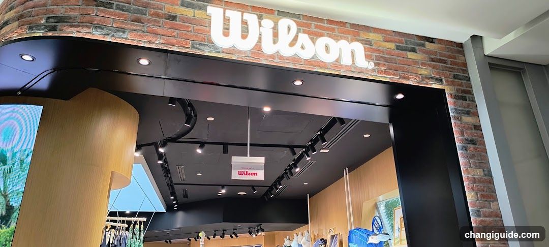 The image shows the exterior entrance of a Wilson store, featuring a brick facade with the brand name 'Wilson' prominently displayed in white lettering above the doorway. The interior is visible through the open entrance, showcasing modern lighting fixtures and wooden paneling.