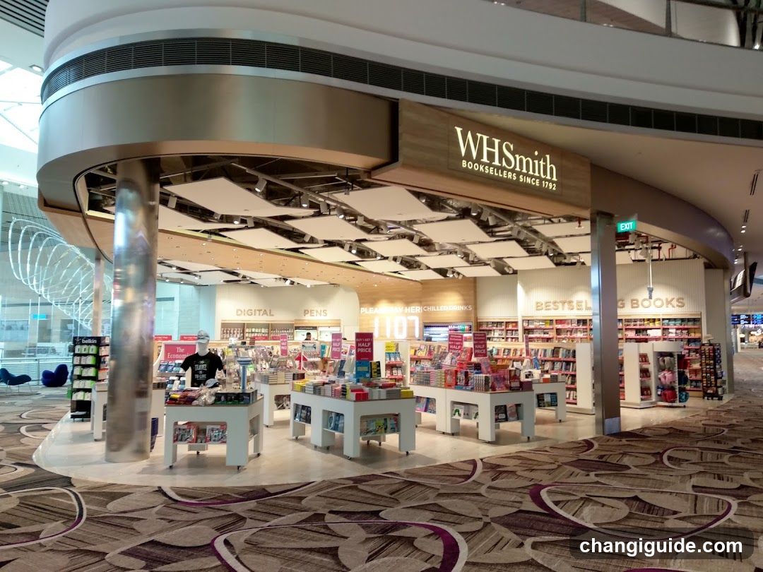 A modern WHSmith bookstore entrance with visible signage reading 'WHSmith BOOKSELLERS SINCE 1972'. The store features a curved facade, illuminated interior displays of books and merchandise, and multiple product sections labeled such as 'DIGITAL', 'PENS', 'BESTSELLING BOOKS', and 'BOOKS'. A staff member is visible inside near the entrance.