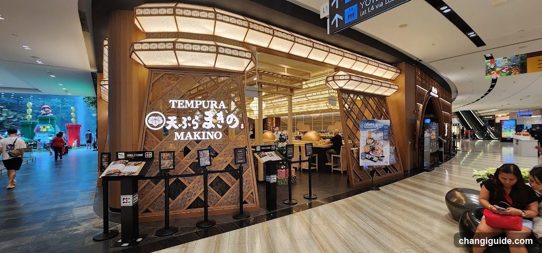 A modern Japanese tempura restaurant with wooden lattice facade and illuminated signage displaying 'TEMPURA MAKINO' in English and Japanese characters. The entrance features black stanchions, menu boards on stands, and a visible interior dining area with warm lighting.