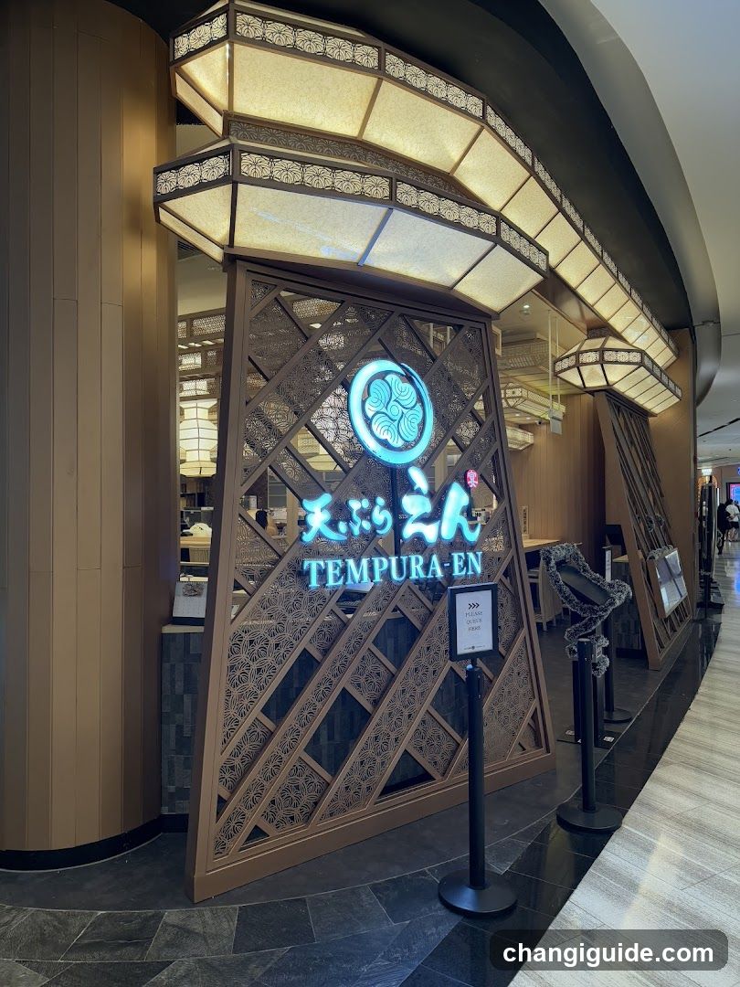 A modern Japanese restaurant entrance featuring a decorative wooden facade with intricate lattice patterns and illuminated signage displaying 'TEMPURA-EN' in both English and Japanese characters. Above the entrance, traditional-style lanterns hang from a curved ceiling structure, casting warm light over the dark tiled floor.