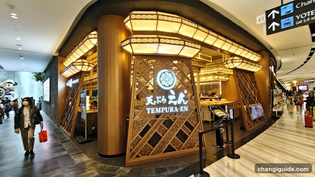 A brightly lit shop front with wooden lattice design and illuminated signage displaying 'TEMPURA-EN' in English and Japanese characters. The entrance features a curved facade with warm lighting, and the interior is partially visible through glass panels.