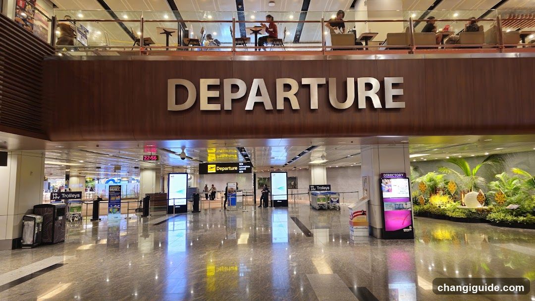 Interior view of an airport departure hall with polished marble floors, a large 'DEPARTURE' sign above the gate area, and a mezzanine level with seating. Visible elements include trolley bins, digital screens, directional signs, and tropical plant displays.