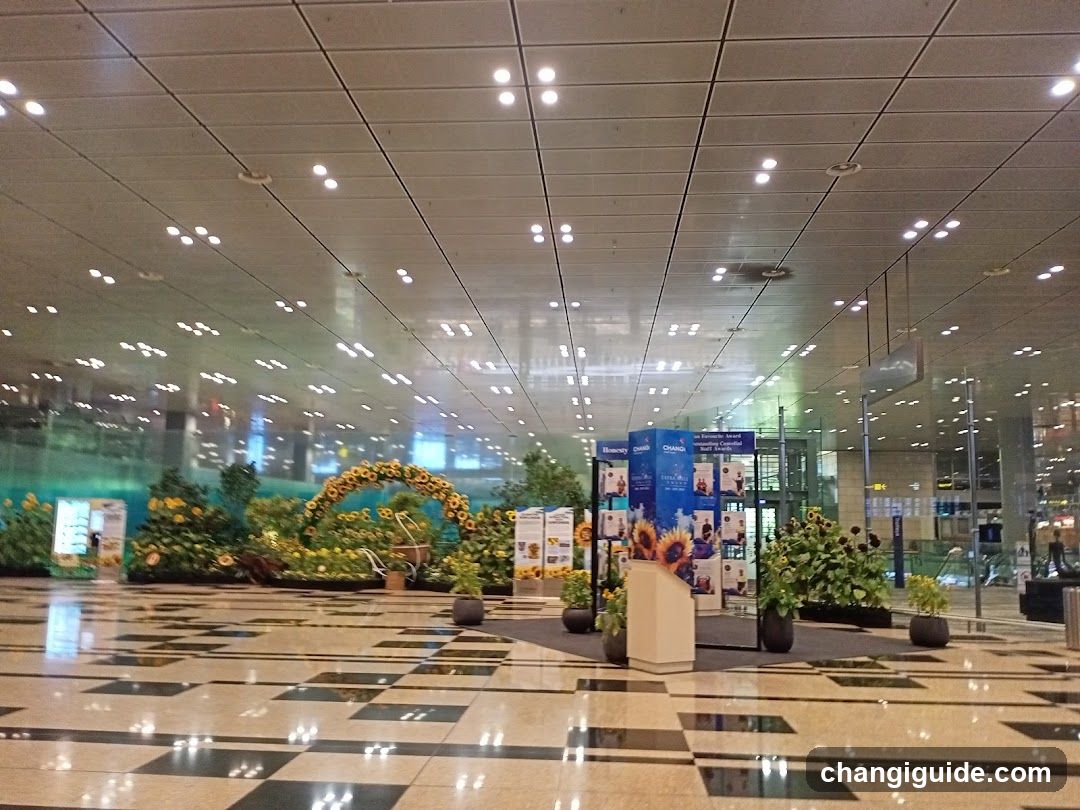 A spacious indoor area with a polished tiled floor and a high ceiling with recessed lighting. The space features decorative elements including sunflower arrangements, greenery, and informational posters. There are no visible shop fronts or menus.