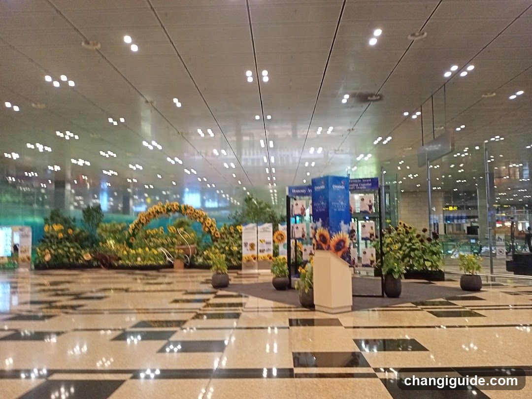 A spacious airport terminal hall featuring polished checkered flooring and a high ceiling with recessed lighting. Large glass windows on the left reveal an outdoor night scene. In the center, there is a promotional display with blue banners showing sunflowers and advertisements. Several potted plants and floral arrangements are placed around the area, including a large arch of yellow flowers in the background.