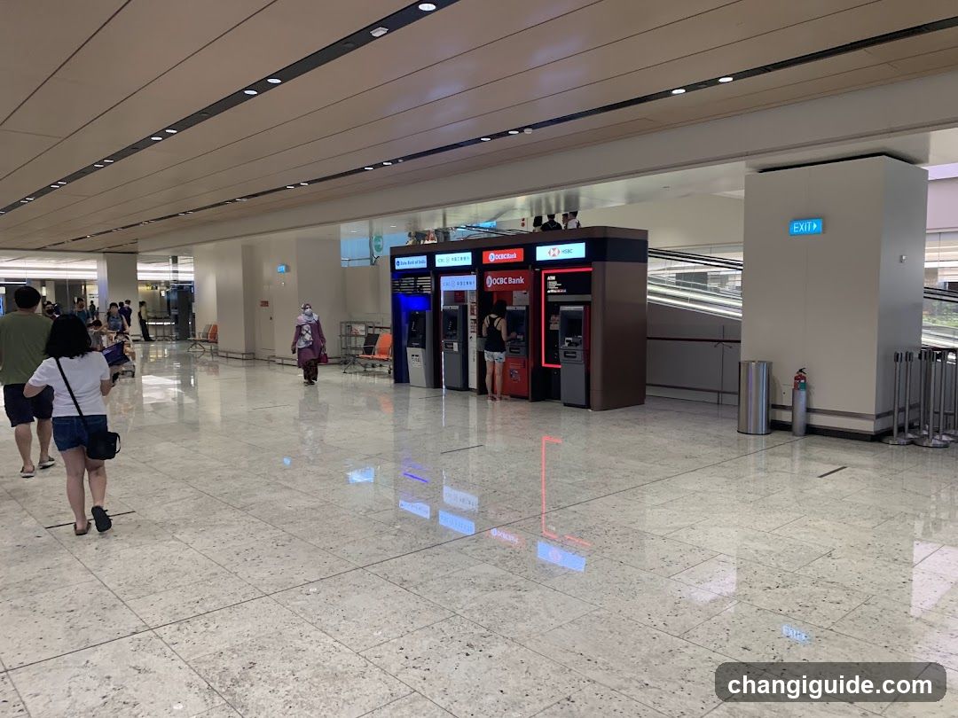 A spacious, well-lit indoor area with polished marble flooring and recessed ceiling lighting. In the center, there is a row of ATMs from OCBC Bank and other banks, including one with red branding. A person in a purple uniform walks nearby, while others are seen in the background near seating areas. An exit sign is visible on the right wall.