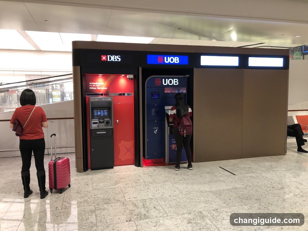 A modern ATM kiosk with DBS and UOB branding, featuring two ATMs under a covered structure. A woman in red stands near the DBS ATM with a pink suitcase, while another person uses the UOB ATM. The setting appears to be an airport terminal with polished floors and visible signage.
