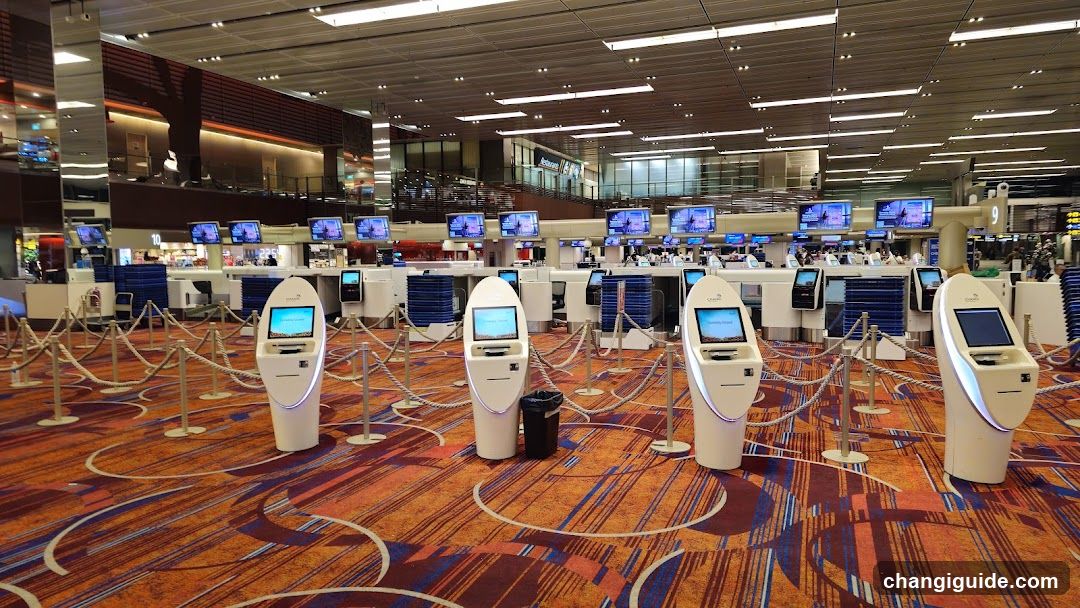 A spacious airport terminal interior featuring multiple self-service kiosk stations arranged in rows on a patterned carpet with orange and blue tones. The kiosks have illuminated screens and are separated by stanchions and ropes. In the background, there is an elevated walkway with glass railings, additional service counters, and digital display boards showing flight information or advertisements. Lighting is bright and modern, with recessed ceiling lights and long fluorescent fixtures.