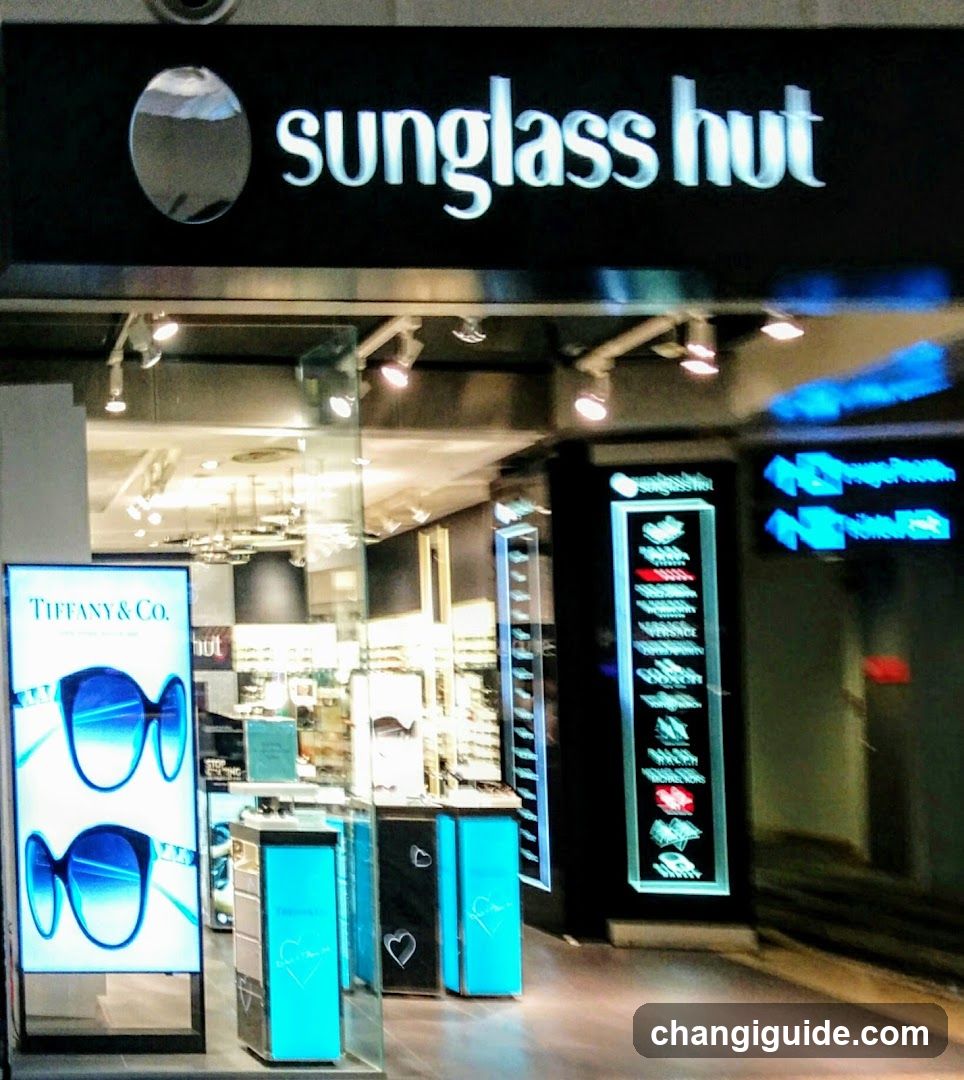 Exterior view of a Sunglass Hut store with illuminated signage and glass entrance. A Tiffany & Co. poster is visible on the left, while an illuminated vertical display showcases sunglasses frames inside the shop.