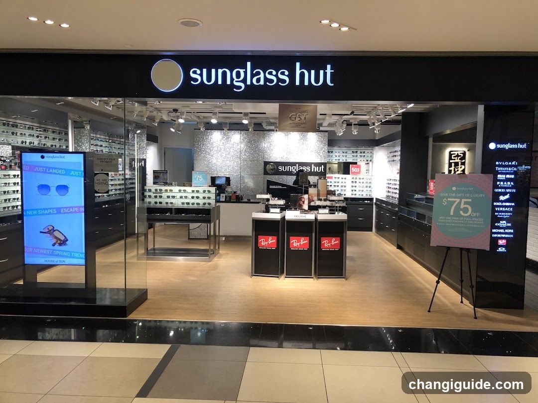 The image shows the storefront of a Sunglass Hut store, featuring prominent signage with the brand name and logo above the entrance. Inside, there are display racks filled with sunglasses, promotional stands showcasing Ray-Ban products, and digital screens advertising new shapes and deals. A sign indicates GST exemption status. The interior is well-lit with modern lighting fixtures.
