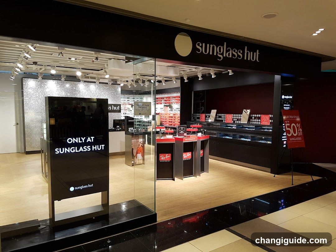 The image shows the exterior of a Sunglass Hut store in a mall. The storefront features large glass windows and doors, with prominent signage displaying 'sunglass hut' on a black background above the entrance. Inside, there are display cases showcasing sunglasses, promotional signs advertising discounts, and a digital screen stating 'ONLY AT SUNGLASS HUT'. The interior is well-lit with track lighting, and the floor is made of light-colored wood or laminate.