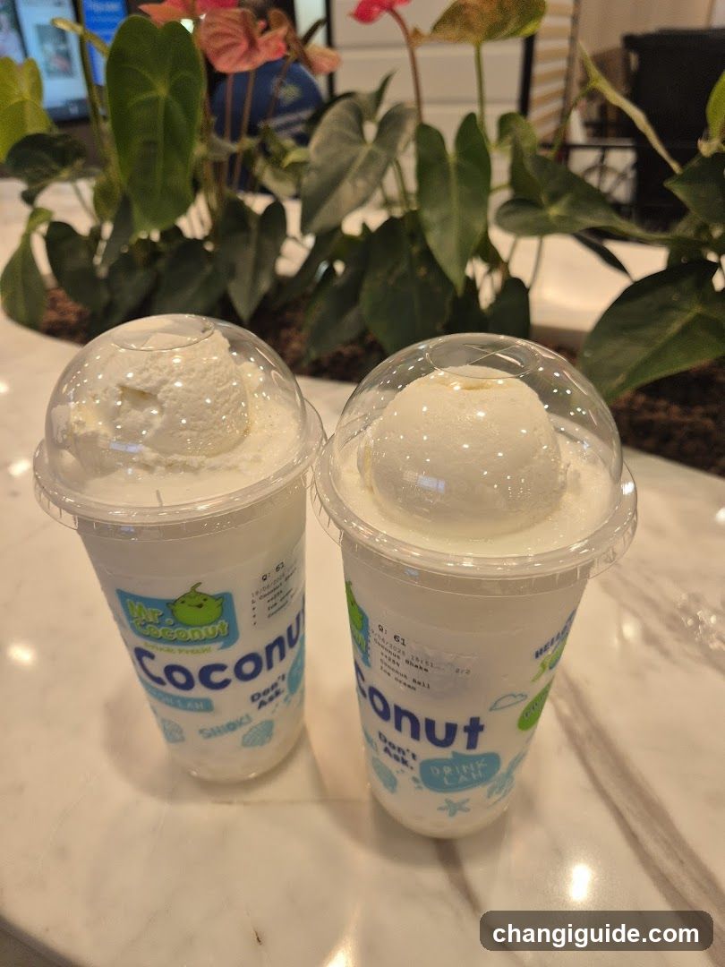 Two cups of coconut-flavored drinks topped with scoops of ice cream, placed on a marble countertop. The cups are branded with 'My Coconut' and feature tropical designs. In the background, there is a potted plant with large green leaves and pink flowers.