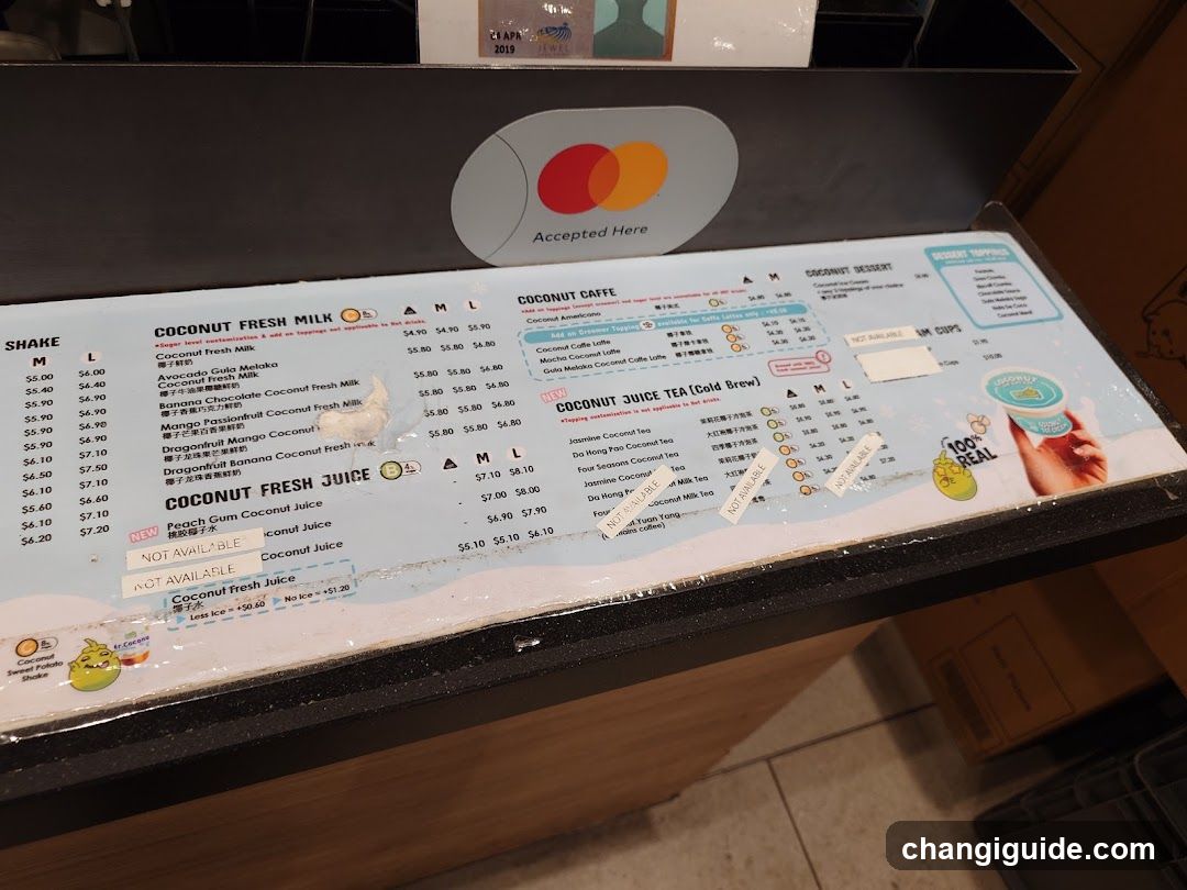 A close-up of a menu board at a shop counter, featuring sections for Coconut Fresh Milk, Coconut Cafe (hot drinks), and Coconut Fresh Juice. The board includes price lists with size options (S, M, L) and some items marked as 'Not Available'. A Mastercard logo is visible above the menu.