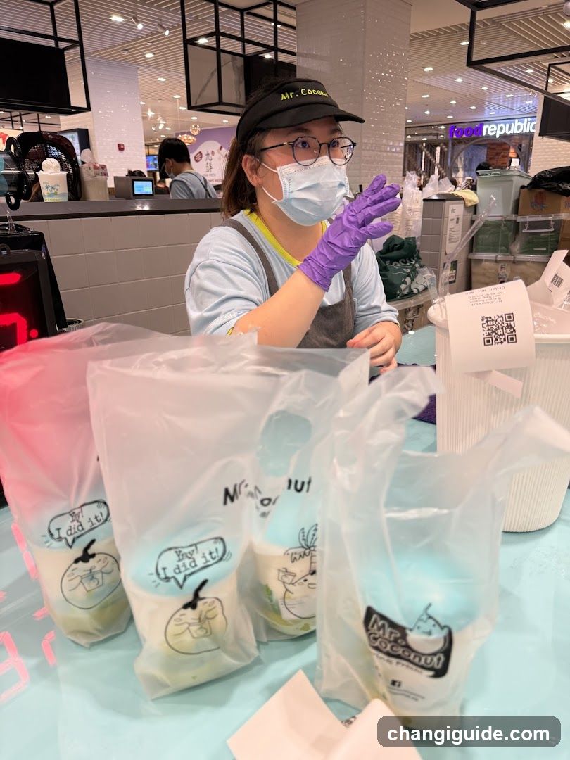 A staff member wearing a mask and gloves is preparing or packaging drinks at a counter in a brightly lit indoor food court setting. Several branded plastic bags with cartoon characters are visible on the counter, along with a QR code sticker attached to a container. The background shows other shops like 'Food Republic' and mall-style ceiling lighting.