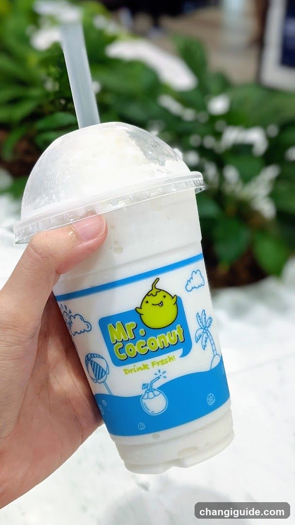 A hand holding a cup of Mr Coconut drink with a straw, featuring a blue and white label with a coconut illustration.