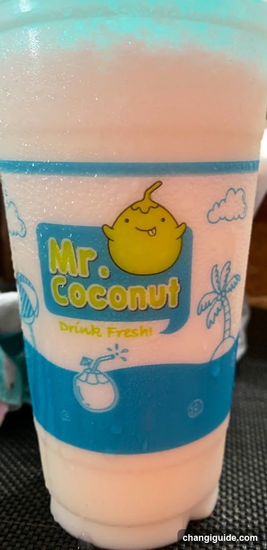 A close-up of a plastic cup containing a creamy, light pink beverage with blue foam on top. The cup features branding for 'Mr. Coconut' with a cartoon coconut character and the slogan 'Drink Fresh!'. Background includes faint illustrations of clouds and palm trees.