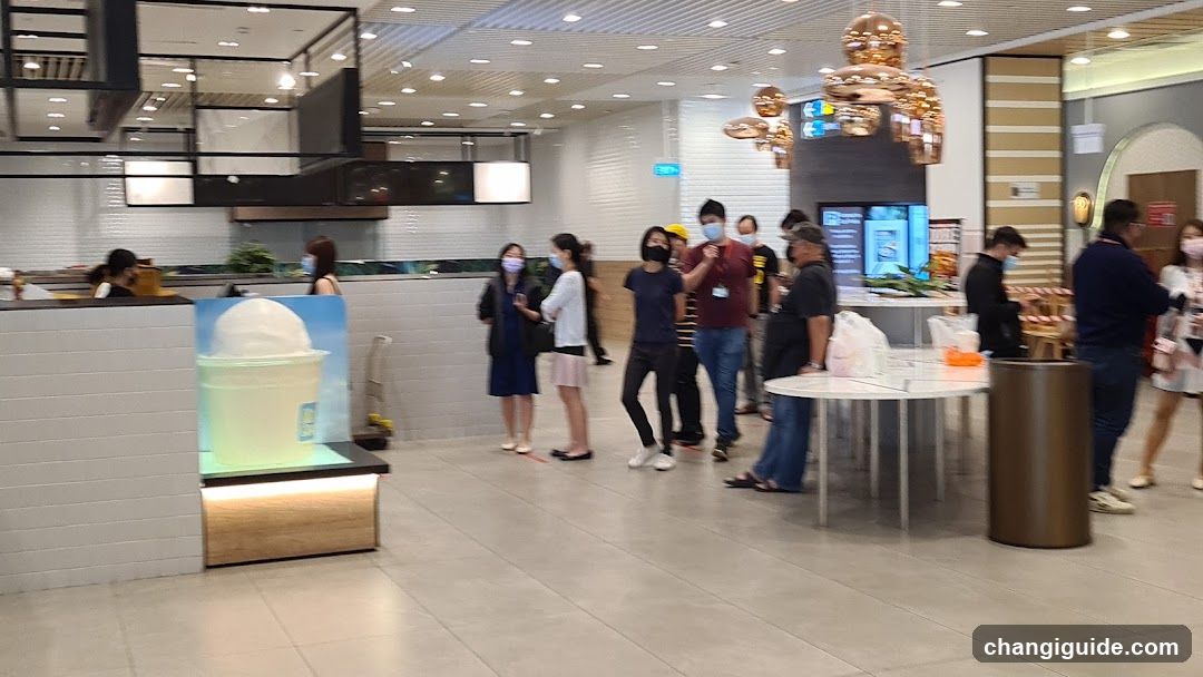 A busy indoor food court area with multiple people standing in line or waiting at counters. Visible elements include a large illuminated display of an ice cream cup on the left, a counter with staff behind it, and several customers wearing masks. A decorative golden sculpture hangs from the ceiling. The background shows kitchen equipment and signage above the counters.