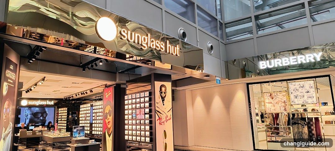 The image shows the exterior of two retail stores in a shopping mall. On the left is Sunglass Hut, featuring illuminated signage with its logo and multiple display windows showcasing eyewear products. To the right is a Burberry store, also with visible branding on its facade. Both storefronts are well-lit and appear to be open for business.