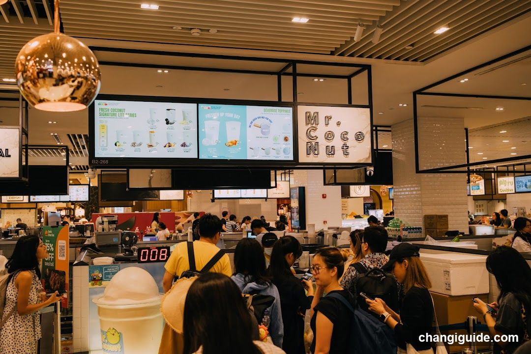 A busy indoor food court scene with customers waiting in line near a counter. A large digital menu board displays drink options and prices above the service area. The shop sign reads 'Mr. Coconut' on the right side of the display. People are standing or sitting, some looking at phones, others facing the counter. Lighting is warm from ceiling fixtures and hanging lamps.