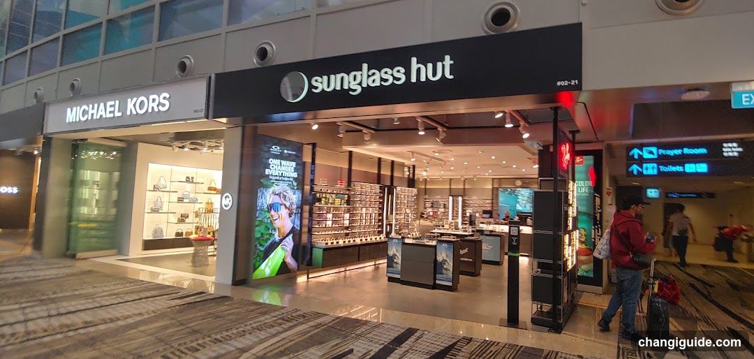 The image shows the exterior of a Sunglass Hut store, with its signage clearly visible above the entrance. The store is located next to a Michael Kors shop. Inside, there are multiple display shelves filled with sunglasses and other accessories. A person is seen walking near the entrance with luggage.