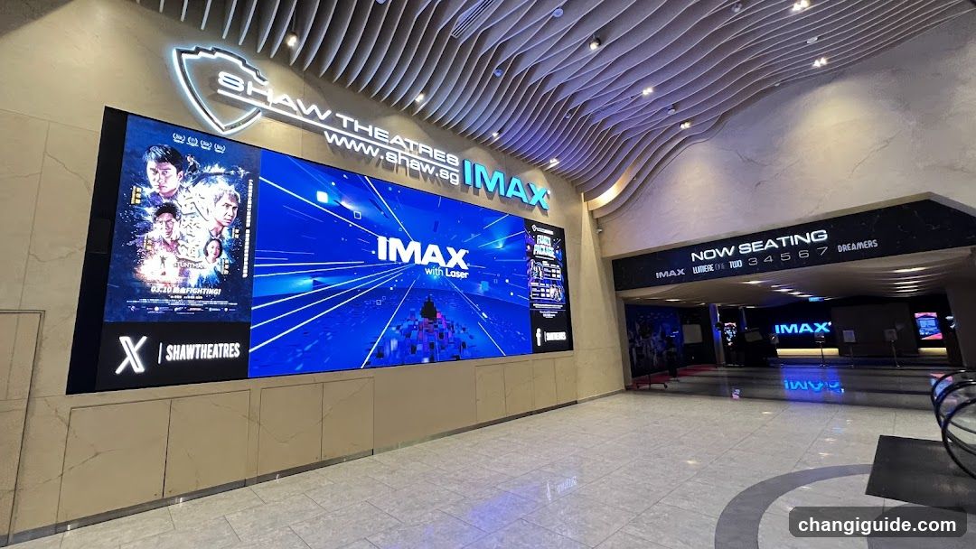 Interior view of a cinema lobby featuring large digital screens displaying movie posters and IMAX branding, with modern ceiling design and signage indicating seating areas.