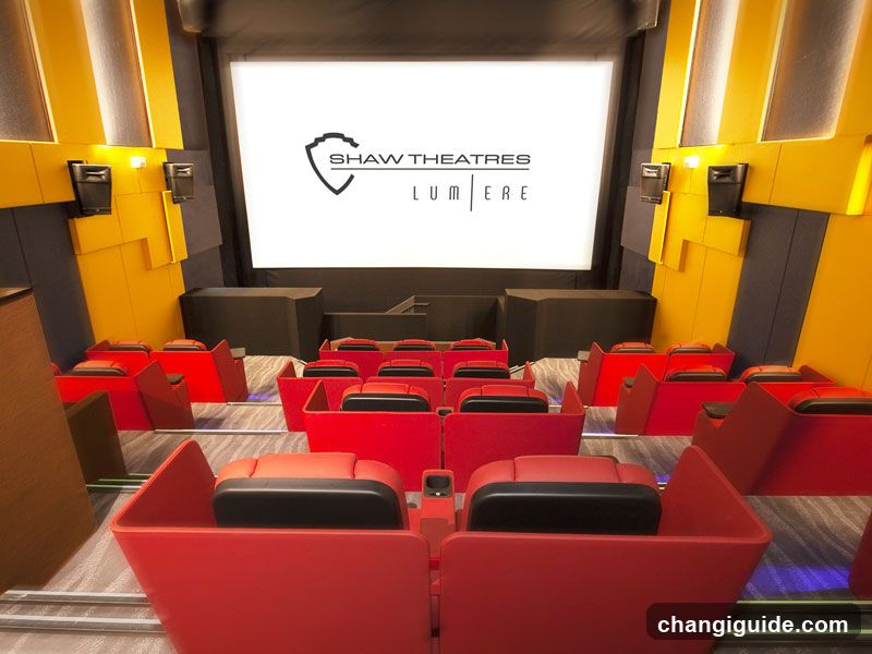 A modern movie theater interior featuring rows of red reclining seats with black headrests, arranged in a tiered layout facing a large white screen displaying the Shaw Theatres Lumiere logo. The walls are accented with yellow and dark gray panels, and ambient lighting enhances the cinematic atmosphere.