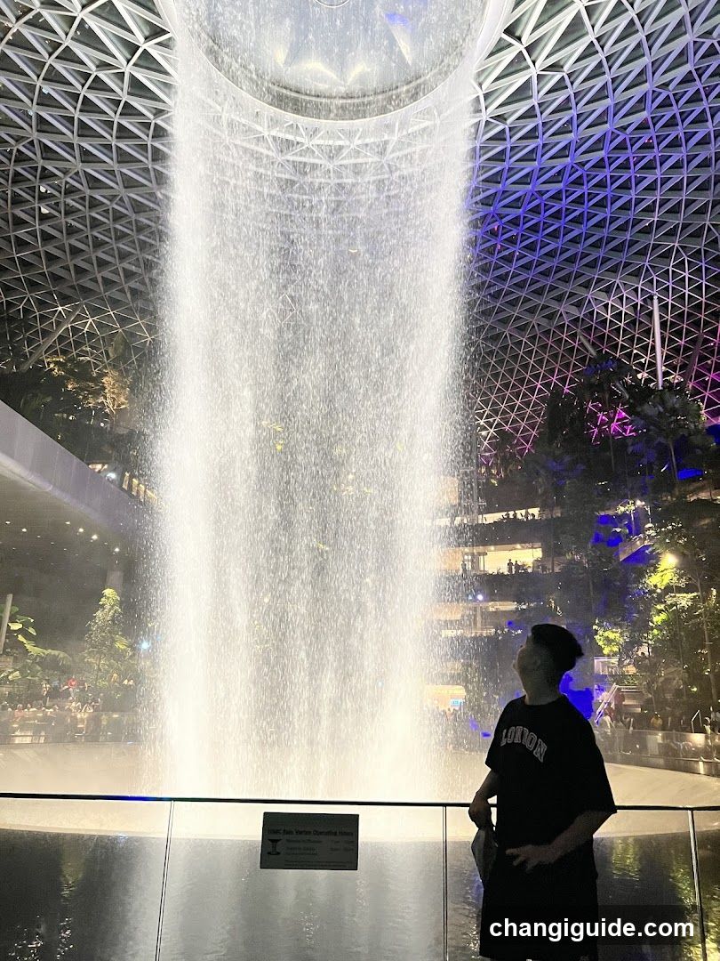 A person stands in front of a large indoor waterfall under a geometric dome ceiling with purple lighting.
