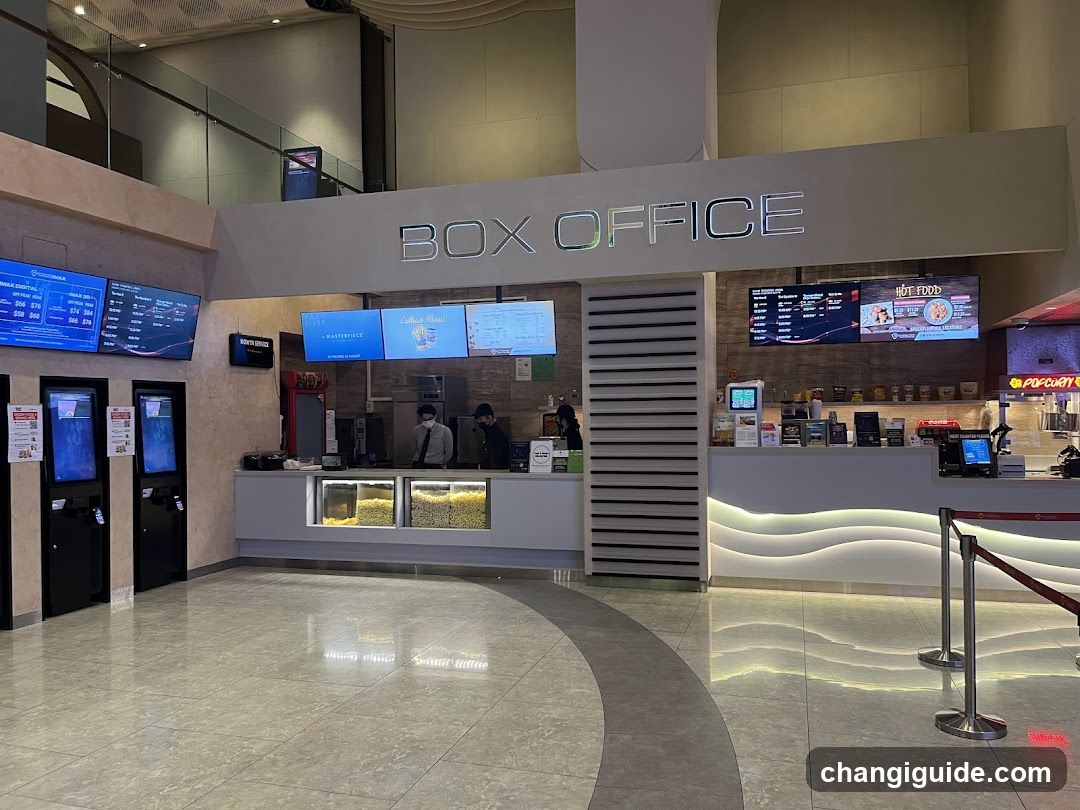 Interior view of a cinema box office with multiple screens displaying movie information, self-service kiosks on the left, and staff behind counters. A coffee shop section is visible to the right.