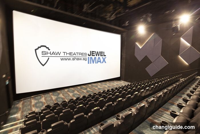 Interior view of a modern cinema auditorium with rows of black reclining seats facing a large white screen displaying the Shaw Theatres Jewel IMAX logo and website URL. Geometric wall panels with backlighting are visible on the right side.