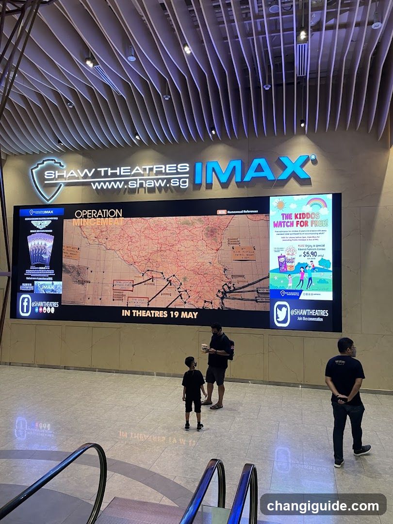 Interior shot of Shaw Theatres IMAX lobby featuring a large digital display showing movie promotion 'Operation Mischief' with release date May 19. Visible signage includes Shaw Theatres logo and website URL. A promotional poster for 'The Kiddos Match For Fun!' is displayed on the right side of the screen. Three people are standing near the bottom, observing or walking past the display. The ceiling has modern wavy wooden paneling with ambient lighting.