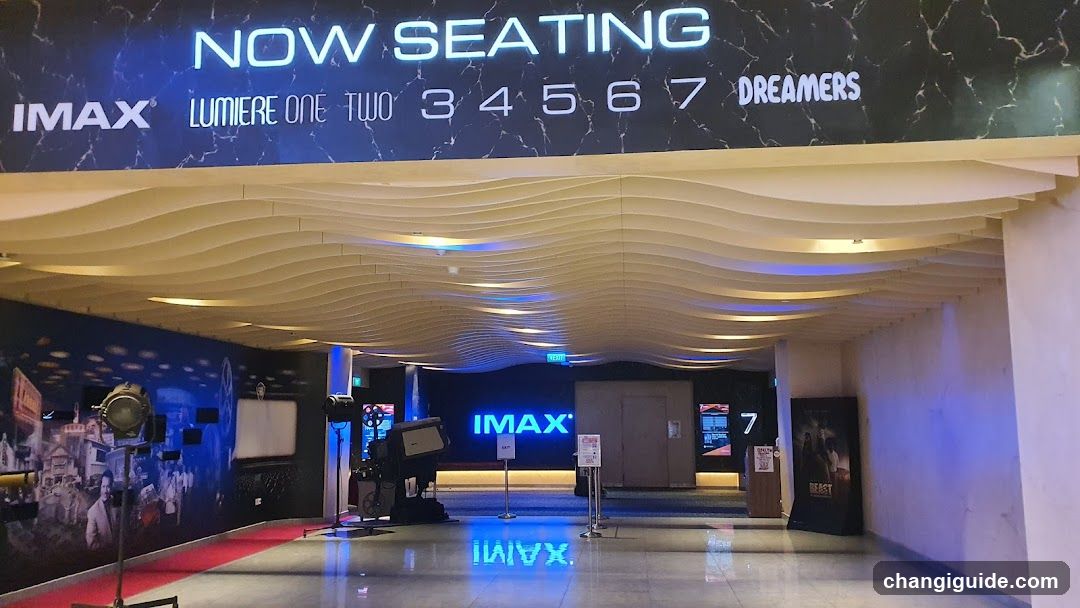Interior view of an IMAX cinema lobby with wavy ceiling design, illuminated signage reading 'NOW SEATING' and seat numbers (34567), IMAX branding on walls and floor reflection, promotional posters including one for the movie 'BEAST', and a red carpet leading to screening area.