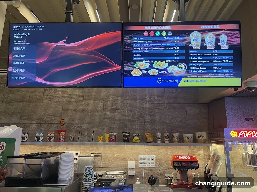 Interior view of a food court at Shaw Theatres Jewel, featuring digital menu boards displaying movie listings and beverage/snack options. Visible items include soft drinks, teas, soups, popcorn, chicken nuggets, and hot wings. A Coca-Cola dispenser is present on the counter below.