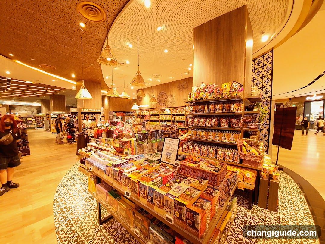 Interior of a brightly lit retail store with wooden accents and hanging pendant lights, featuring shelves stocked with packaged goods, snacks, and merchandise displayed on tiered stands.
