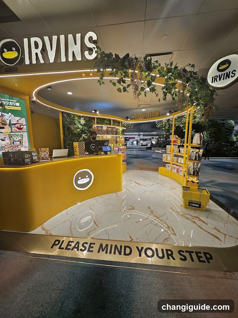 The image shows the interior of an Irving's store, featuring a bright yellow counter with the brand logo and a curved display area filled with various products. The floor is marble-like with a 'PLEASE MIND YOUR STEP' warning at the entrance. Greenery decorates the ceiling, adding a natural touch to the modern design.
