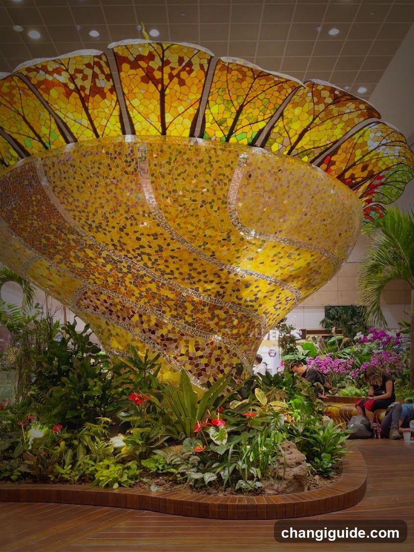 A large, vibrant mosaic sculpture resembling a tree or flower with golden-yellow and orange hues dominates the scene. The structure is adorned with intricate patterns and surrounded by lush greenery and colorful flowers. People are seated nearby, enjoying the display.