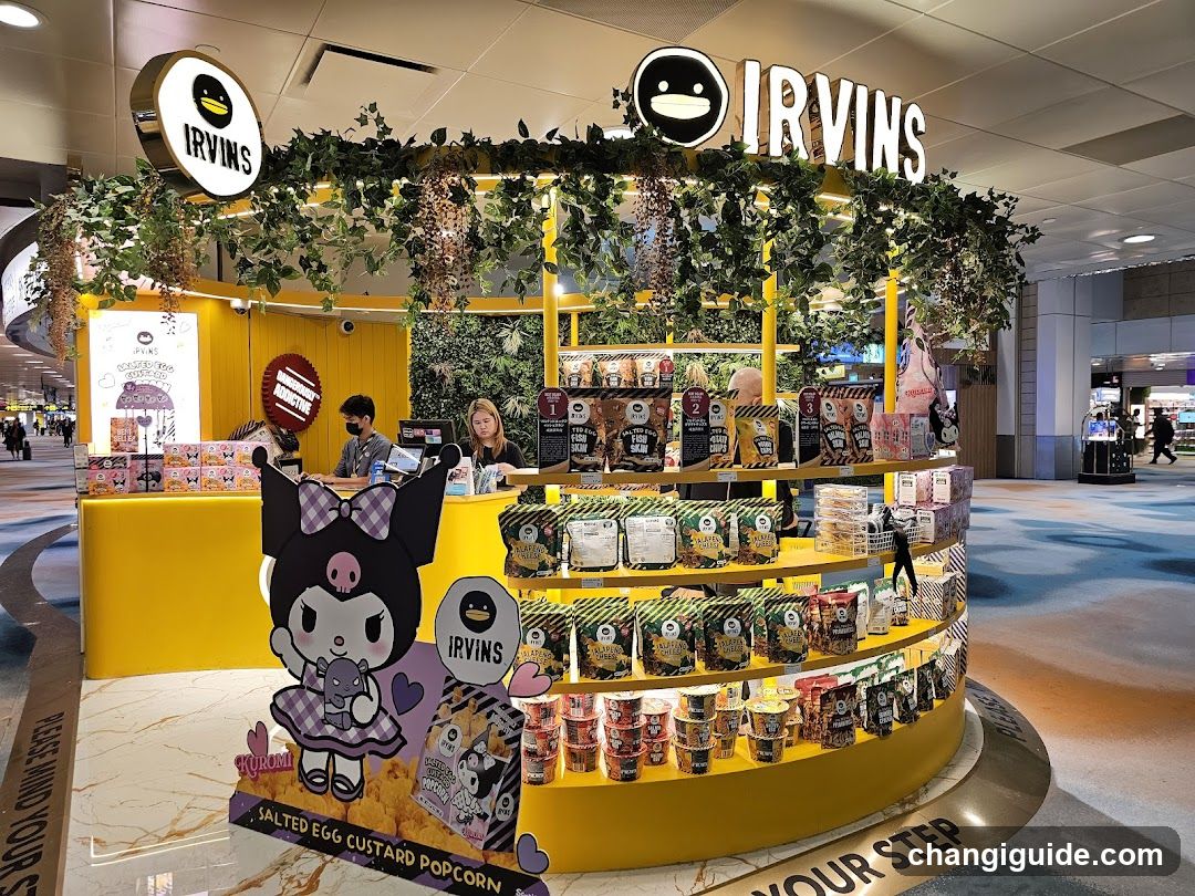 A brightly lit yellow kiosk with greenery and hanging plants, featuring IRVINS branding on signage above. A large Kuromi character cutout stands in front. Shelves display packaged popcorn and cup noodles. Staff are visible behind the counter.