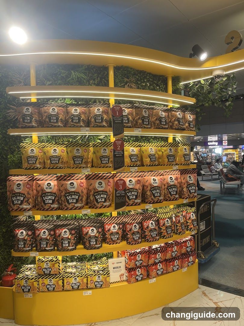 A brightly lit yellow curved display shelf stocked with various packaged snacks, including salmon skin and other savory items. The shelves are adorned with greenery and illuminated by LED strips. In the background, an airport terminal is visible with passengers seated on benches.