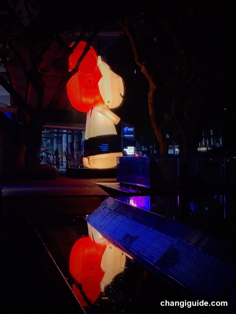 A nighttime outdoor scene featuring a brightly lit, stylized sculpture resembling a figure in red and white, standing near a water feature that reflects its glow. In the background, there is a building entrance with visible signage reading 'Sakura', though no menu or detailed storefront details are present.