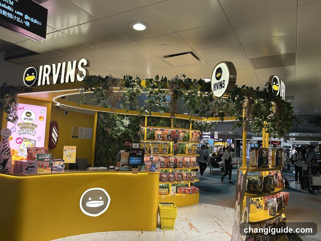 A brightly lit interior of an Irving's popcorn shop, featuring yellow counters and shelves stocked with various snack products. Greenery decorates the ceiling area above the counter. A digital menu board is visible on the left wall displaying colorful graphics and text in Japanese characters.