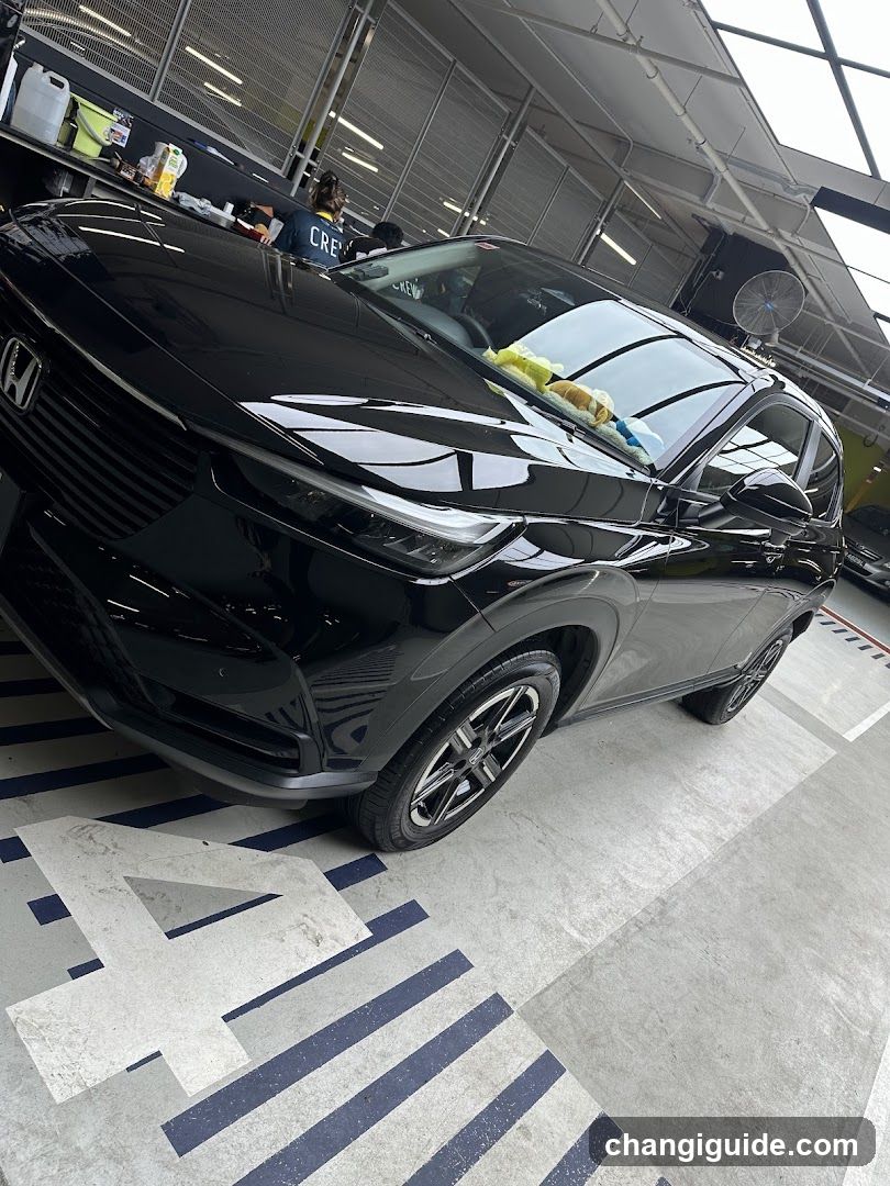 A black Honda HR-V is parked inside a detailing or car wash facility. The vehicle's glossy finish reflects the overhead lights and surroundings. A yellow microfiber cloth rests on the windshield, suggesting recent or ongoing cleaning. In the background, another person wearing a 'CREW' shirt works near a counter with various bottles of detailing products. The floor features blue and white striped markings, typical of automotive service bays.