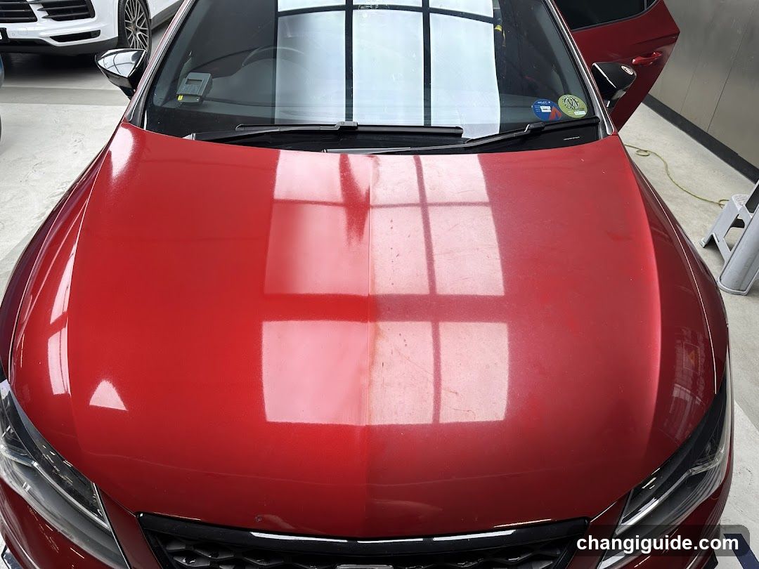 A close-up view of a red car's hood, showcasing reflections from overhead lights and windows in what appears to be an indoor showroom or garage setting.
