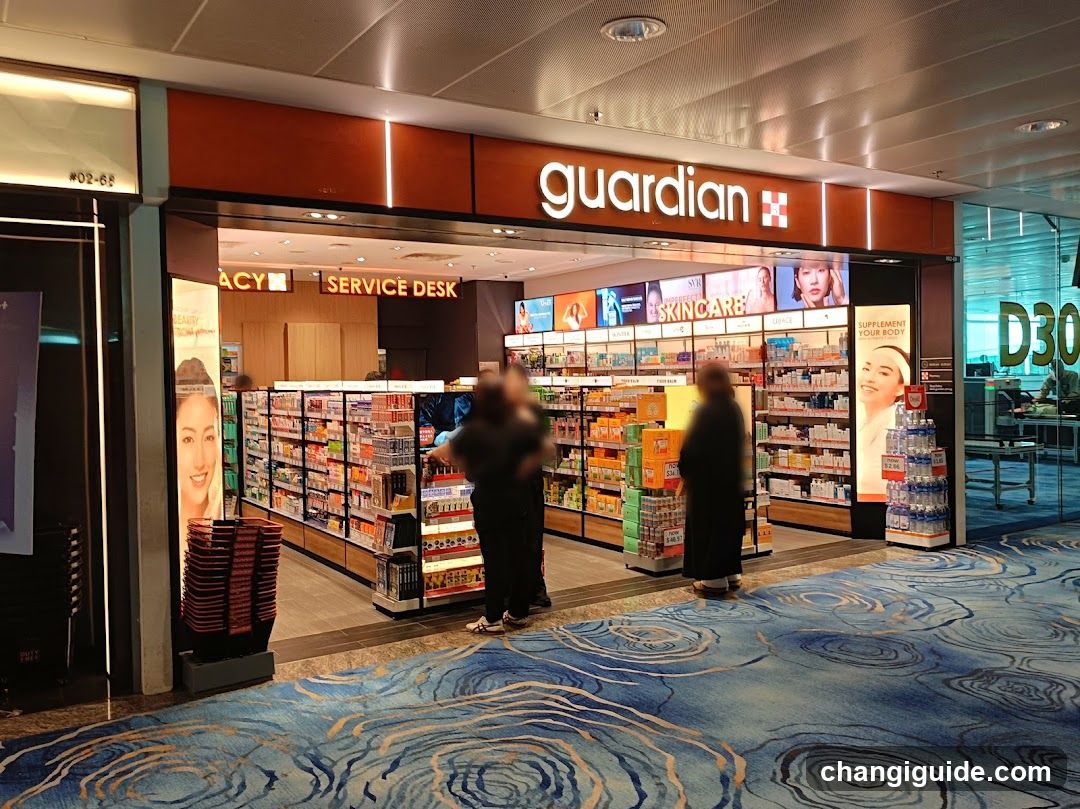 The photo shows the storefront of a Guardian convenience store, with its illuminated signage prominently displayed above the entrance. Inside, shelves are stocked with various products including skincare items and beverages. A service desk is visible in the background, along with promotional posters on the walls. The floor features a patterned blue carpet.