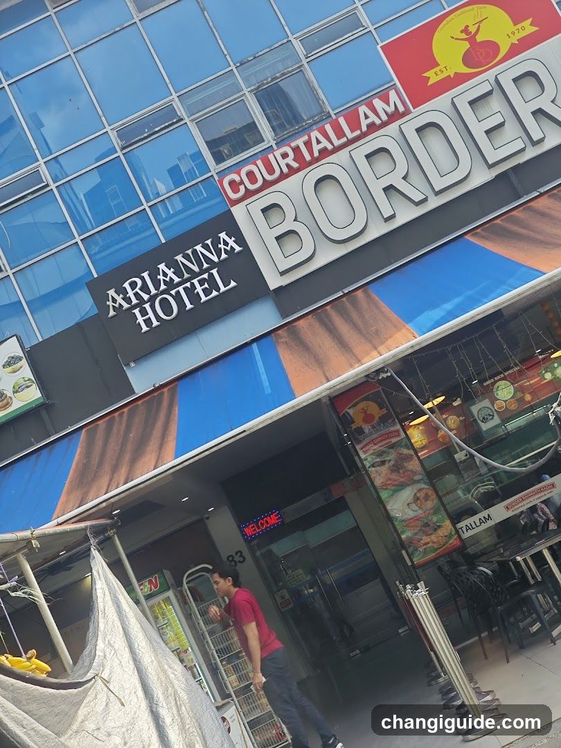 Exterior view of a commercial building featuring signage for 'Arlanna Hotel' and 'Cour Tallam Border'. The facade includes blue-tinted glass windows, a striped awning in blue and brown tones, and an entrance with glass doors displaying a neon 'WELCOME' sign. A person in red is walking near the entrance. Outdoor seating is partially visible on the right side.