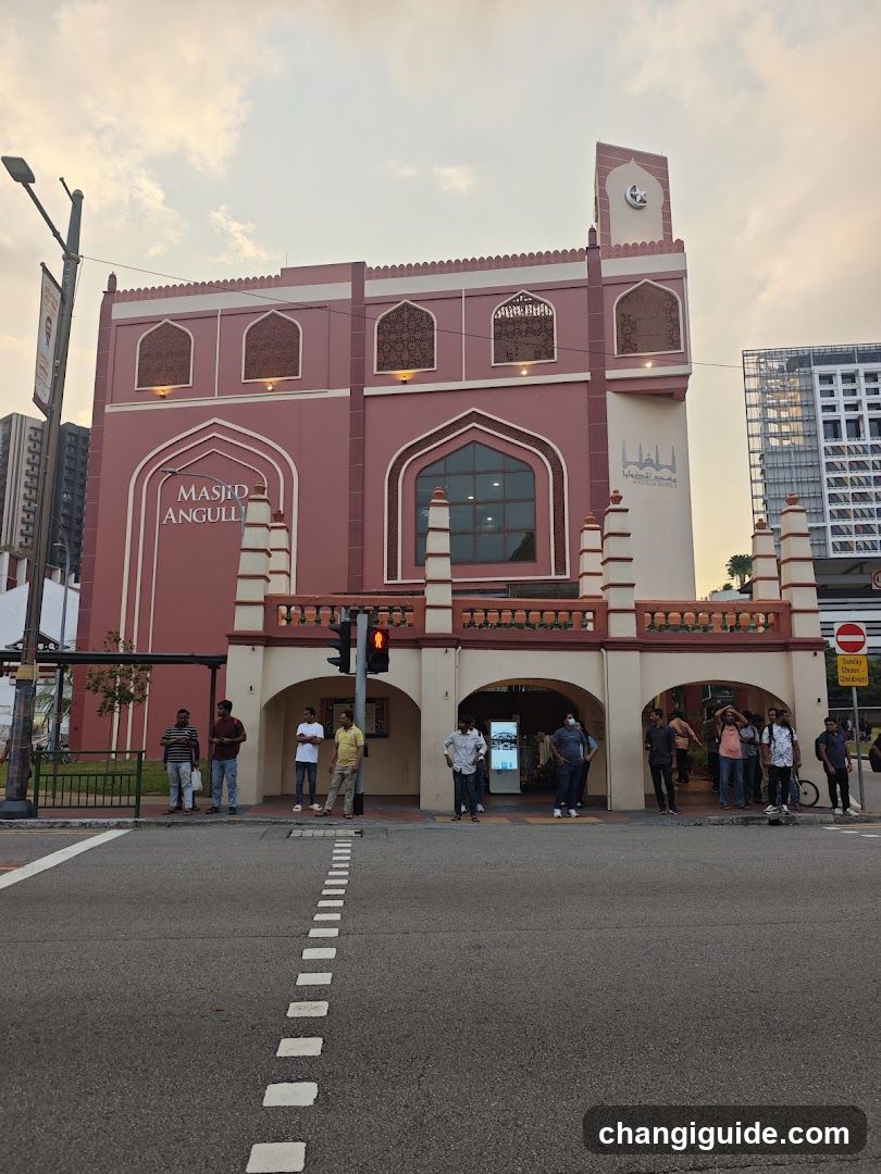 A pink building with arched windows and a sign reading 'MASJID ANGULIN' on the facade. People are gathered near the entrance.