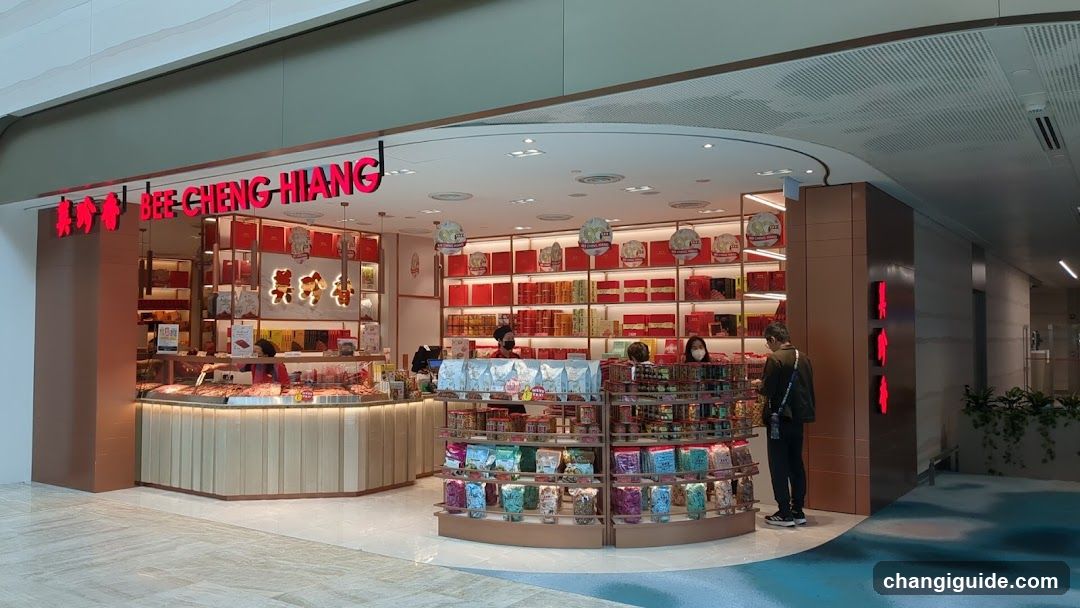 The image shows the storefront of a Bee Cheng Hiang shop, featuring illuminated signage with both Chinese and English text. The entrance is wide, with shelves stocked with red-packaged products visible inside. A display counter with various food items is present at the front, along with hanging lanterns. Staff members are seen behind the counter.
