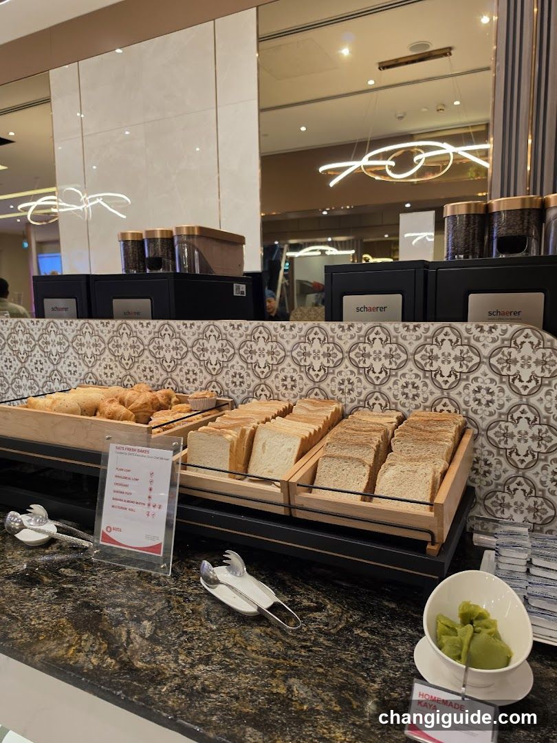 A buffet-style food display featuring trays of sliced breads, croissants, and a bowl of green spread labeled 'Homemade Kaya'. The counter has a decorative tile backsplash with geometric patterns. A sign lists bread options including white, whole wheat, and rye varieties. Coffee dispensers are visible in the background.